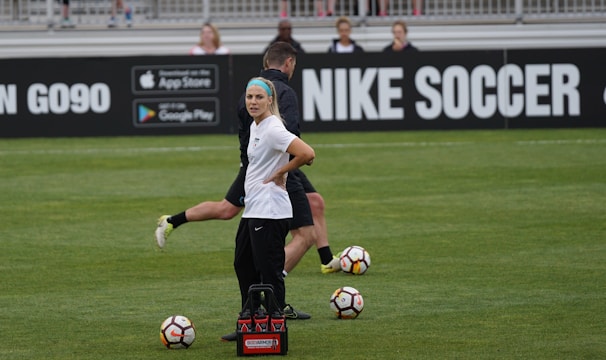 A person in sports attire stands on a soccer field, surrounded by several soccer balls and a carrier with bottles. The field is bordered by advertisements for Nike Soccer and app stores. Several people sit in bleachers in the background.