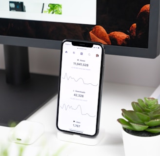A smartphone displays analytics data with graphs showing views, downloads, and likes. It is resting vertically on a white dock next to a small potted succulent plant. In the background, a large computer monitor is partially visible, along with a closed laptop and a white wireless earbud case.