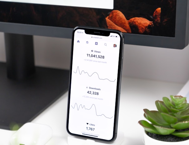A smartphone displays analytics data with graphs showing views, downloads, and likes. It is resting vertically on a white dock next to a small potted succulent plant. In the background, a large computer monitor is partially visible, along with a closed laptop and a white wireless earbud case.