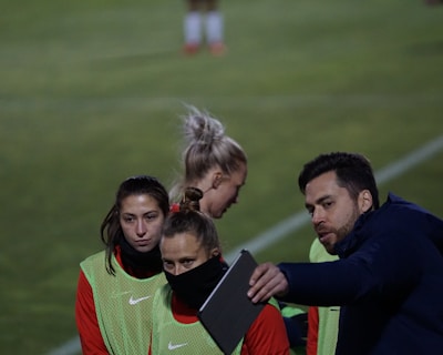A group of individuals on a sports field are wearing green vests over red attire. One person is holding a tablet and seems to be discussing or showing something to the others. The atmosphere suggests a sports event, likely soccer, indicated by their clothing and the setting. The background is a grassy field.