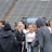 A group of people, likely athletes, are gathered together on a sports field. They are wearing black jackets with some individuals dressed in white sports uniforms underneath. A few people have their hair tied up in buns, and most are engaged in conversation. The background shows tiered stadium seats, suggesting the setting is an outdoor sports venue.