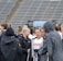 A group of people, likely athletes, are gathered together on a sports field. They are wearing black jackets with some individuals dressed in white sports uniforms underneath. A few people have their hair tied up in buns, and most are engaged in conversation. The background shows tiered stadium seats, suggesting the setting is an outdoor sports venue.