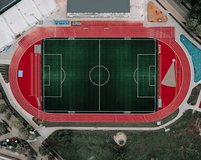 An aerial view of a large sports complex featuring a central green football field surrounded by a red running track. Adjacent to the track, there is a parking area, buildings, and pathways. Nearby, there are additional sports courts in different colors, including a blue swimming pool and orange basketball courts.