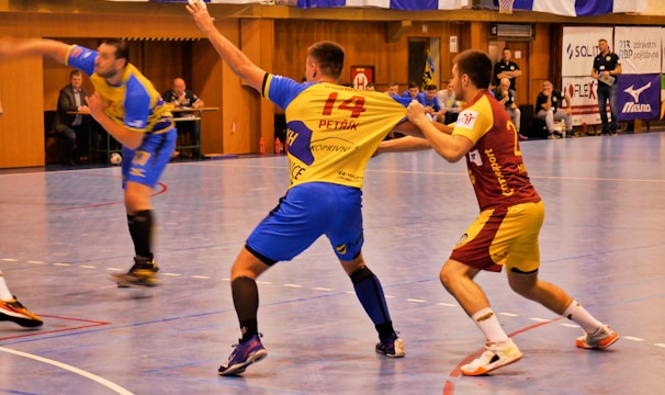 A dynamic indoor sports scene with players in motion on a court. Two athletes in blue and yellow uniforms are engaged in an intense handball game, with one player gripping the other's jersey. The background shows other players, several seated people, and advertisements on the walls.
