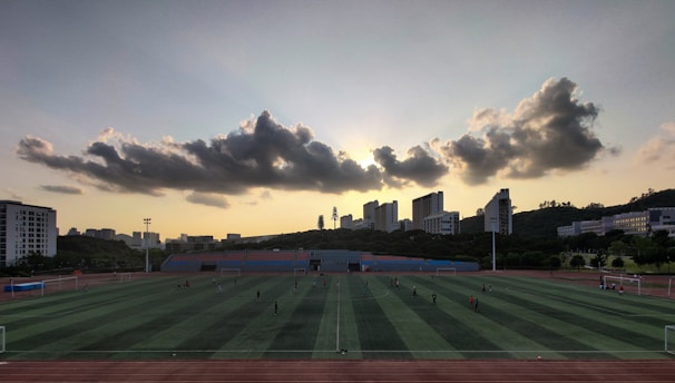 A sports field is situated in the foreground, with several people engaged in activities. The field is surrounded by a running track, and there are goalposts on either end. Behind the field, a line of modern buildings is visible, partially covering a hillside. Above, a dramatic cloud formation stretches across the sky, with the sun just below the horizon creating a silhouette effect.