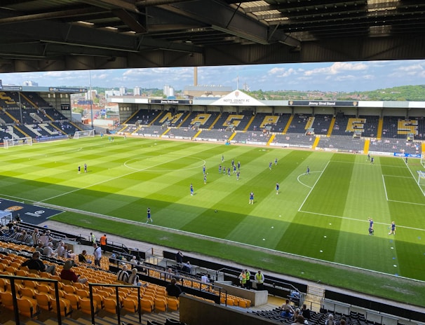 A sports stadium with a well-maintained grass field is depicted. Players are spread across the field, engaging in warm-up activities. The stands are partially filled with spectators, and the stadium is surrounded by urban buildings under a partly cloudy sky.