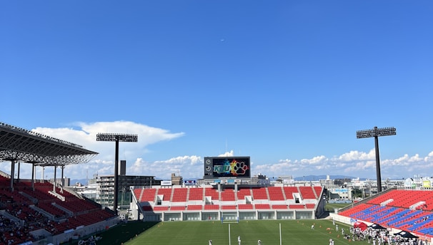 A large sports stadium with red and blue seating overlooks a green field with a goalpost at the center. A few people are scattered across the field, with some tents and a gathering of individuals on the right side. The sky is clear and blue with some clouds visible in the distance. A large screen or scoreboard is situated at the far end of the field.